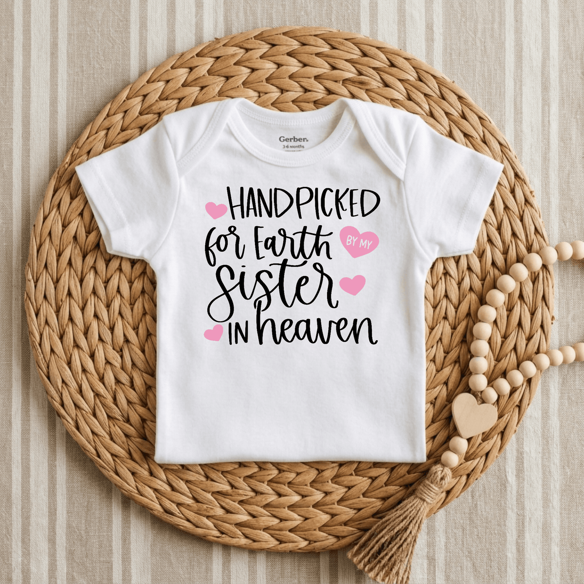 The Toodleroos Handpicked for Earth by My Sister in Heaven Baby Onesie is displayed on a woven mat, featuring black script reading “Handpicked for Earth by My Sister in Heaven,” with pink heart accents and a wooden bead garland beside it. This sibling remembrance bodysuit provides a meaningful way to honor your baby’s special guardian angel sister, making it a heartfelt memorial keepsake from Toodleroos.