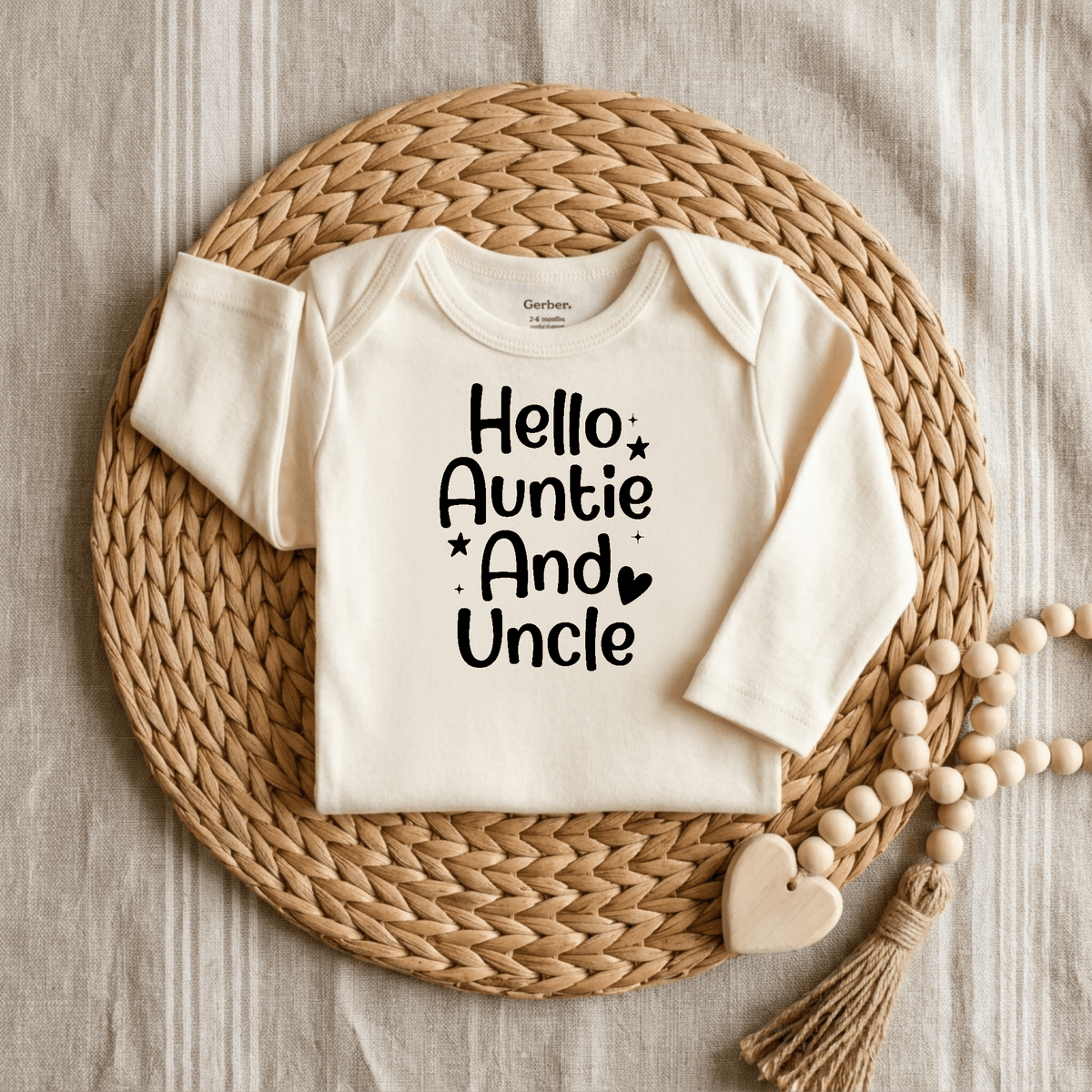 The Toodleroos Hello Auntie and Uncle Onesie – Baby Announcement Bodysuit features bold black lettering on a cream baby onesie, beautifully displayed on a woven mat with a wooden beaded garland and heart accent atop a striped fabric background.