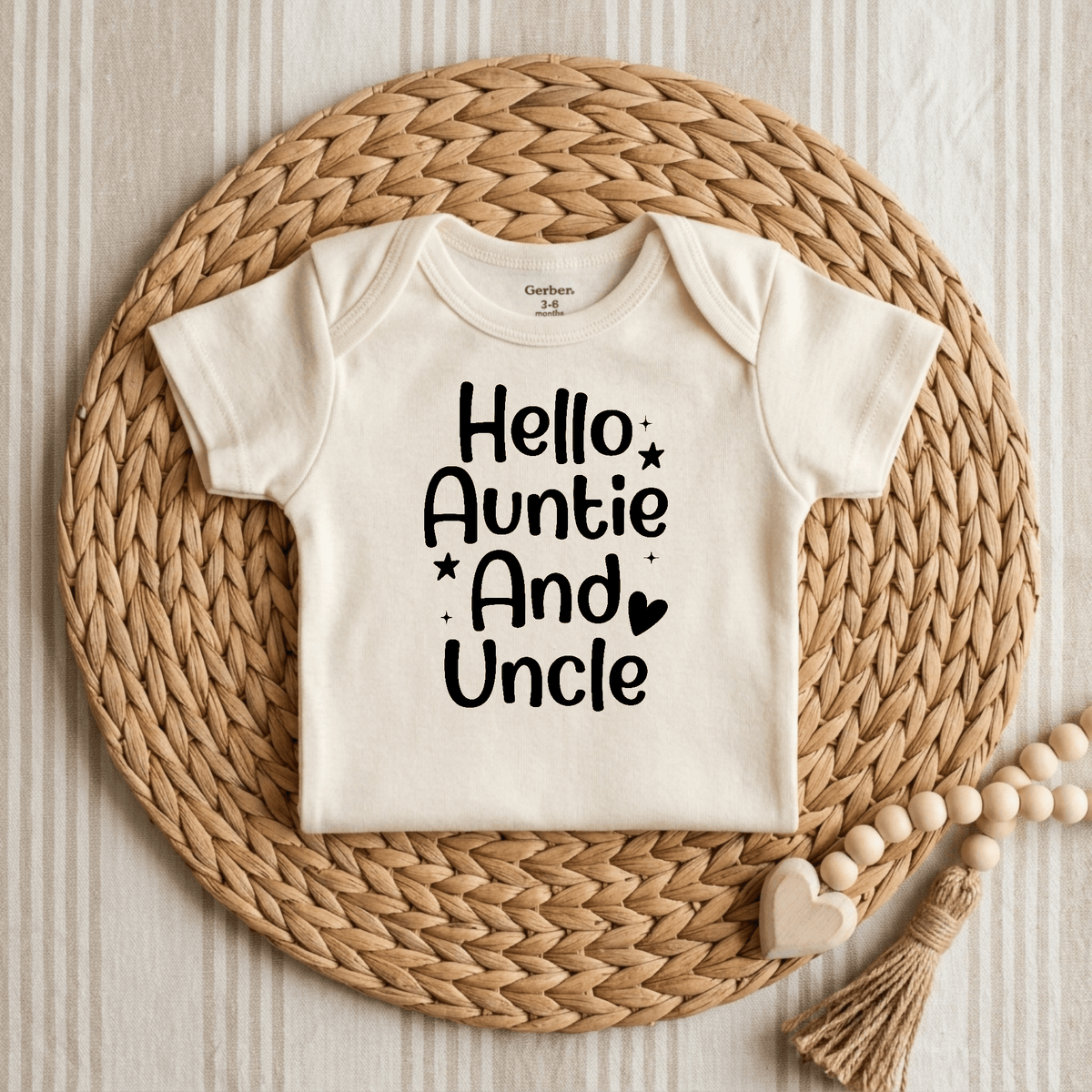 A cream baby onesie from Toodleroos, displayed on a woven mat, features the words “Hello Auntie and Uncle” in black lettering along with small stars and a heart. This Hello Auntie and Uncle Onesie – Baby Announcement Bodysuit is perfect for a special pregnancy reveal. A wooden bead and heart decoration is placed nearby on the mat.