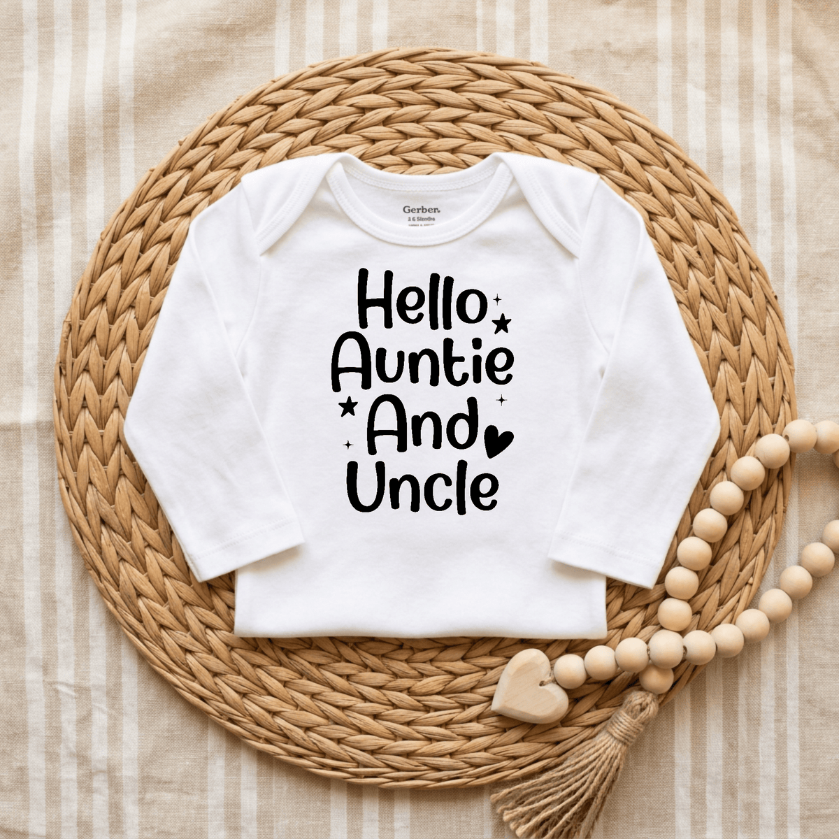 The Toodleroos “Hello Auntie and Uncle Onesie – Baby Announcement Bodysuit” features the phrase “Hello Auntie And Uncle” in black on a white onesie, making it perfect for pregnancy reveals or announcements. It is showcased on a woven placemat with a wooden beaded garland and heart decoration nearby.