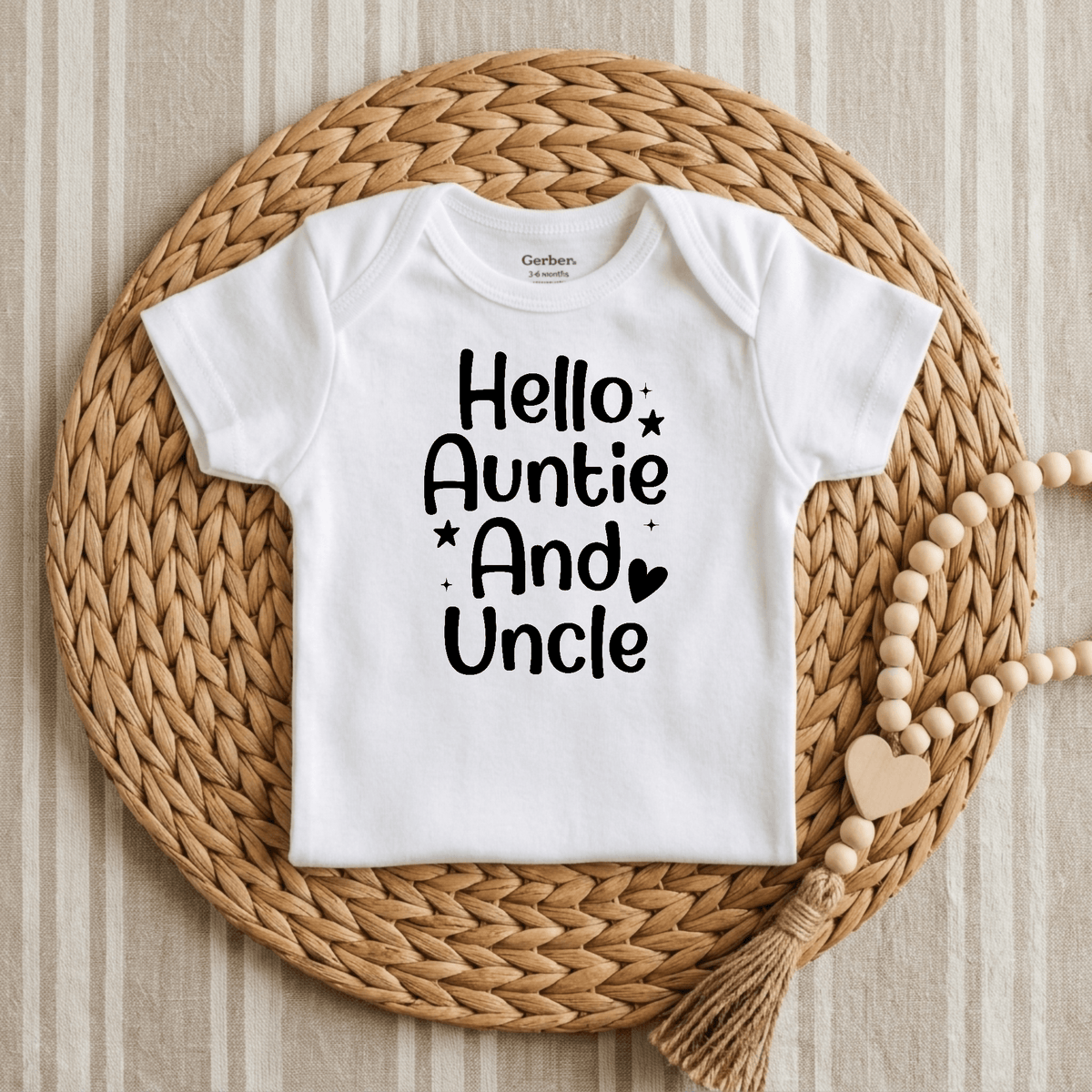 The Toodleroos Hello Auntie and Uncle Onesie – Baby Announcement Bodysuit features black “Hello Auntie And Uncle” lettering on a white baby onesie, making it perfect as an aunt and uncle reveal or pregnancy announcement. It’s pictured laid on a round woven mat with a beaded tassel decoration beside it, set against a striped fabric background.