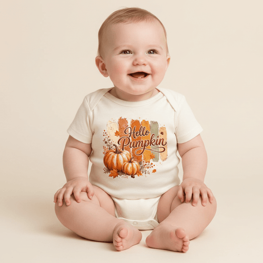 A smiling baby wears the Toodleroos Hello Pumpkin Tee – a cozy white fall shirt for babies, toddlers, and youth, adorned with orange pumpkins and playful “Hello Pumpkin” text—perfect for family photoshoots on a light backdrop.
