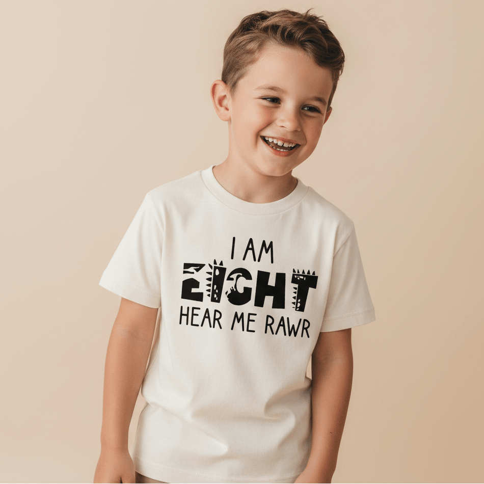 A smiling young boy with brown hair wears the Toodleroos “I Am Eight Hear Me Rawr” Kids Shirt – a dinosaur-themed 8th birthday tee featuring playful animal print lettering. He stands against a plain beige background.