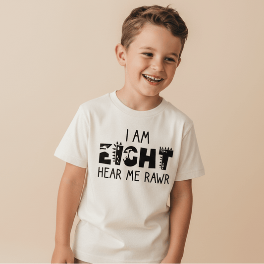 A smiling young boy with brown hair wears the Toodleroos “I Am Eight Hear Me Rawr” Kids Shirt – a dinosaur-themed 8th birthday tee featuring playful animal print lettering. He stands against a plain beige background.