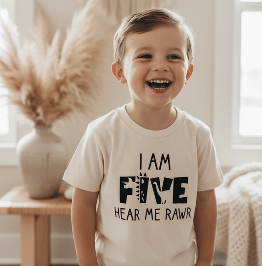 A smiling young boy stands indoors near a vase with pampas grass, wearing the Toodleroos “I Am Five Hear Me Rawr” Kids Shirt – a fun dinosaur 5th birthday tee.