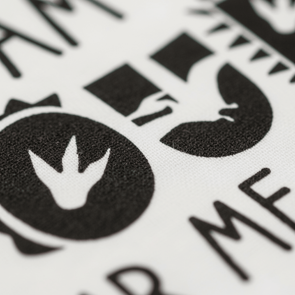 Close-up of black dinosaur-themed symbols and text printed on white fabric from the Toodleroos “I Am Four Hear Me Rawr Kids Shirt – Dinosaur 4th Birthday Tee,” featuring a dinosaur footprint inside a circle and partial bold letters—perfect for celebrating a child’s 4th birthday with a fun dino shirt.