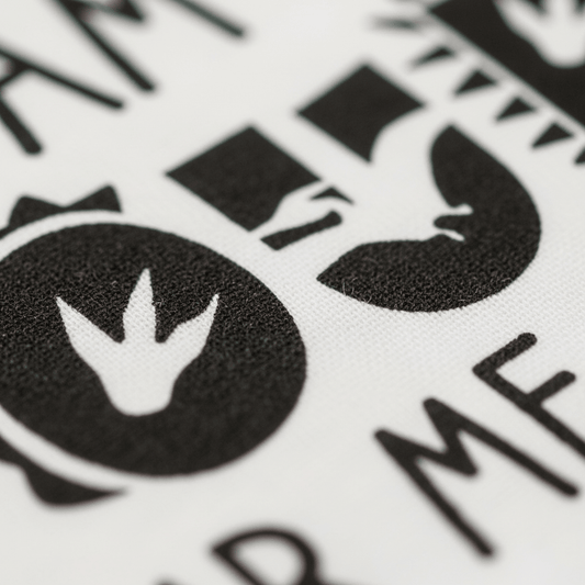 Close-up of black dinosaur-themed symbols and text printed on white fabric from the Toodleroos “I Am Four Hear Me Rawr Kids Shirt – Dinosaur 4th Birthday Tee,” featuring a dinosaur footprint inside a circle and partial bold letters—perfect for celebrating a child’s 4th birthday with a fun dino shirt.