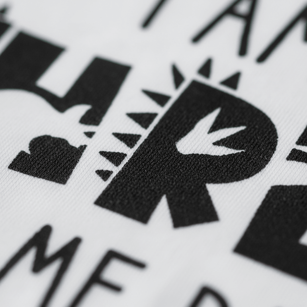 Close-up of black text on white fabric from the Toodleroos “I Am Three Hear Me Rawr” Toddler Shirt—Dinosaur 3rd Birthday Tee, featuring the letter R creatively designed with a dinosaur footprint and spikes to enhance its prehistoric, dino-themed look. Some surrounding letters and shapes are partially visible. Perfect as a dinosaur birthday shirt for toddlers turning three.