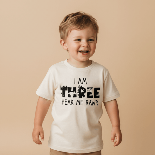A smiling young child wears the Toodleroos “I Am Three Hear Me Rawr Toddler Shirt – Dinosaur 3rd Birthday Tee,” featuring playful dinosaur-themed lettering, while standing against a beige background.