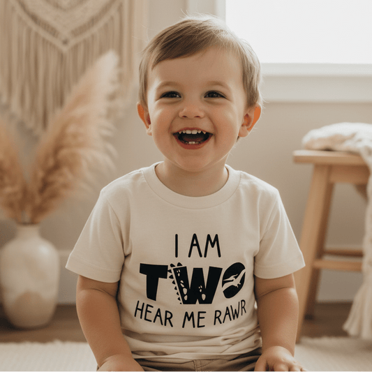 A cheerful toddler wears the Toodleroos “I Am Two Hear Me Rawr” Dinosaur 2nd Birthday Tee indoors, surrounded by neutral décor like a vase with pampas grass and a wooden stool—making this playful toddler shirt perfect for a second birthday or cake smash outfit.