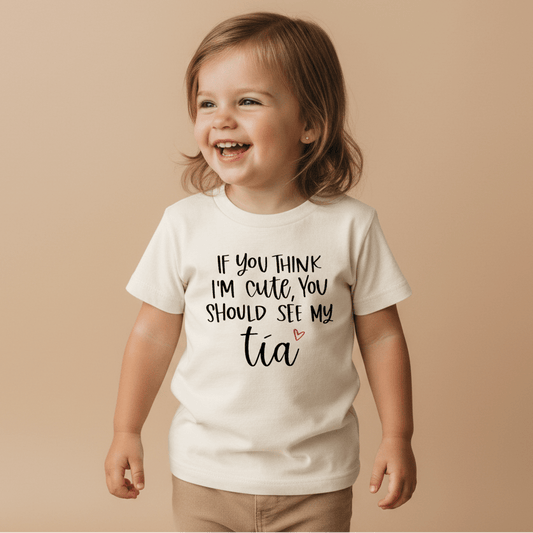 A happy toddler models the Toodleroos “If You Think I’m Cute See My Tía” Toddler Shirt – a funny bilingual tee with a playful message and small pink heart, set against a beige background—a delightful gift for any adorable niece or nephew and their proud aunt.