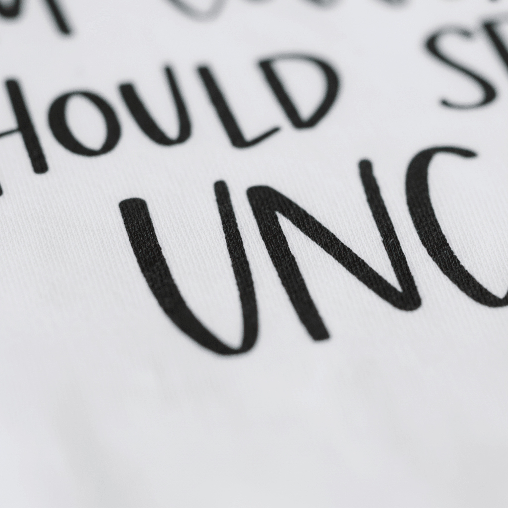 Close-up of a white fabric featuring bold, black hand-lettered text. Only part of the phrase is visible, revealing the words SHOULD and part of UNC—suggesting the playful design of the “If You Think I’m Cute See My Uncle Baby Onesie” by Toodleroos, a funny uncle-themed bodysuit that makes an adorable baby shower gift.
