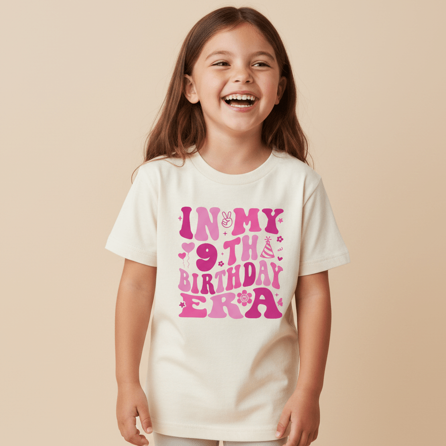 A smiling young girl wears the Toodleroos In My 9th Birthday Era Kids Shirt—a retro 9th birthday tee featuring playful pink lettering and party-themed icons—making it the perfect trendy shirt for celebrating her special day.