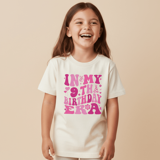 A smiling young girl wears the Toodleroos In My 9th Birthday Era Kids Shirt—a retro 9th birthday tee featuring playful pink lettering and party-themed icons—making it the perfect trendy shirt for celebrating her special day.