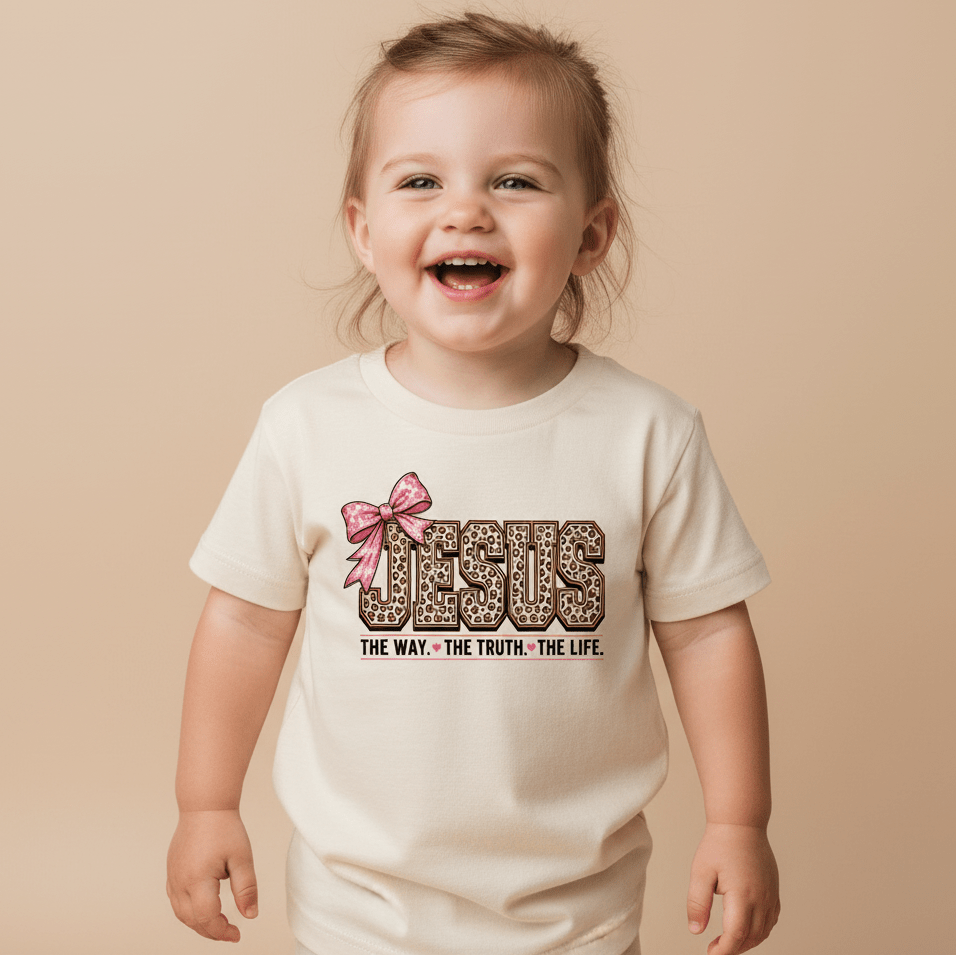 A smiling toddler wears the Toodleroos Jesus The Way The Truth The Life Toddler Girls Shirt, a cream leopard print faith tee featuring a pink bow graphic and the text “Jesus. The Way. The Truth. The Life.” on the front, standing against a beige background.