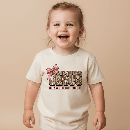 A smiling toddler wears the Toodleroos Jesus The Way The Truth The Life Toddler Girls Shirt, a cream leopard print faith tee featuring a pink bow graphic and the text “Jesus. The Way. The Truth. The Life.” on the front, standing against a beige background.