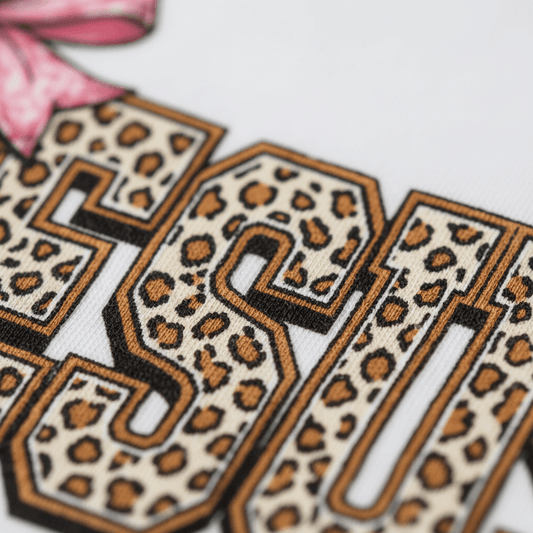 Close-up of large “Jesus The Way The Truth The Life” letters in a leopard print pattern on fabric, partially visible with a pink ribbon in the corner against a white background—perfect for a stylish faith-based toddler girls shirt or trendy leopard print tee from Toodleroos.
