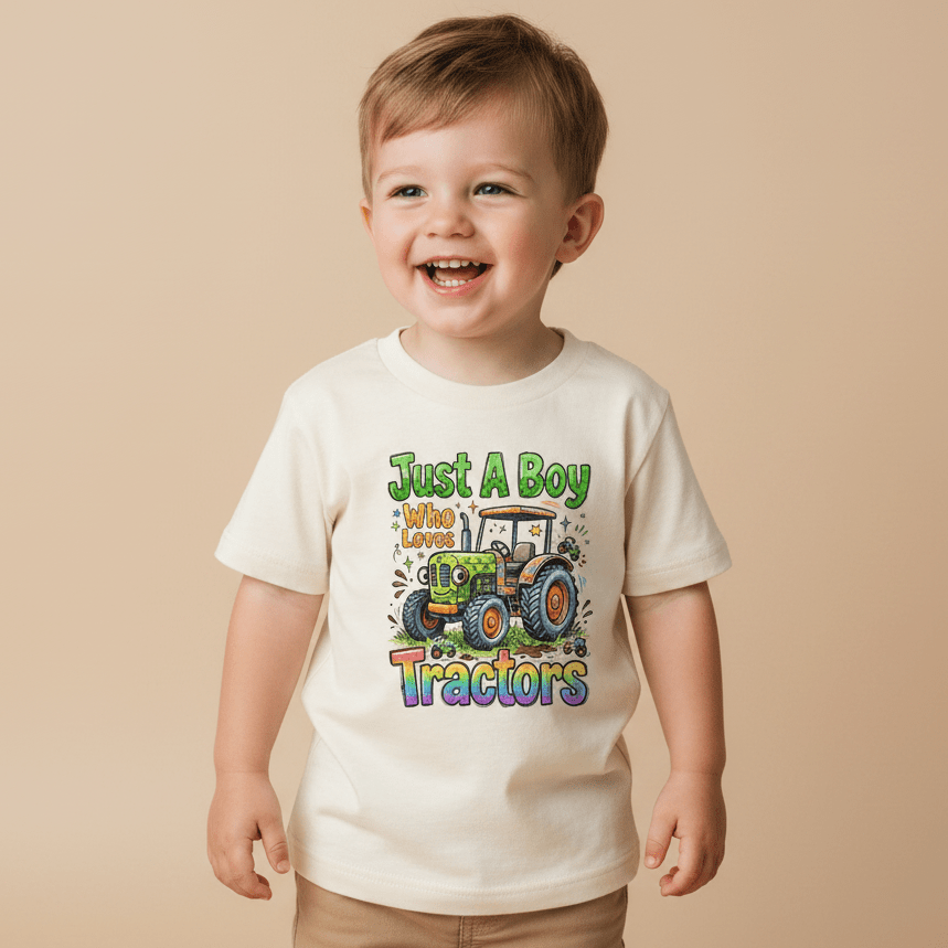 A smiling young boy wears the Toodleroos Just a Boy Who Loves Tractors Toddler Shirt – Farm Tractor Tee, featuring a cream color, cartoon tractor graphic, and playful text, standing against a beige background.