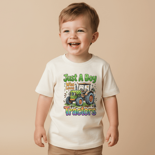 A smiling young boy wears the Toodleroos Just a Boy Who Loves Tractors Toddler Shirt – Farm Tractor Tee, featuring a cream color, cartoon tractor graphic, and playful text, standing against a beige background.