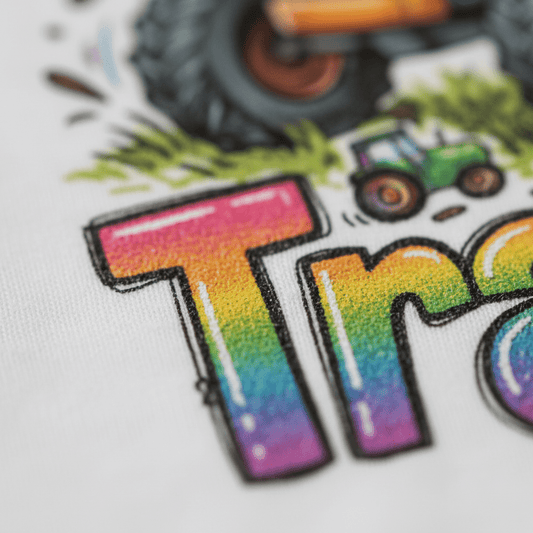 Showcasing a close-up of vibrant, gradient letters on soft fabric, the Toodleroos “Just a Boy Who Loves Tractors” Toddler Shirt features whimsical grass accents and a cheerful green tractor above the playful text. A large black tire peeks at the top, adding extra charm to this farm tractor tee—perfect for any little adventurer who loves spending days outdoors.