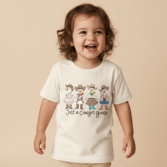 A happy toddler wears the Toodleroos “Just a Cowgirl Goose Toddler Girls Shirt – Western Tee,” a white top featuring five cartoon geese dressed as cowgirls and the playful phrase “Just a Cowgirl Goose” on the front—an adorable western-style shirt perfect for little ones with country charm.