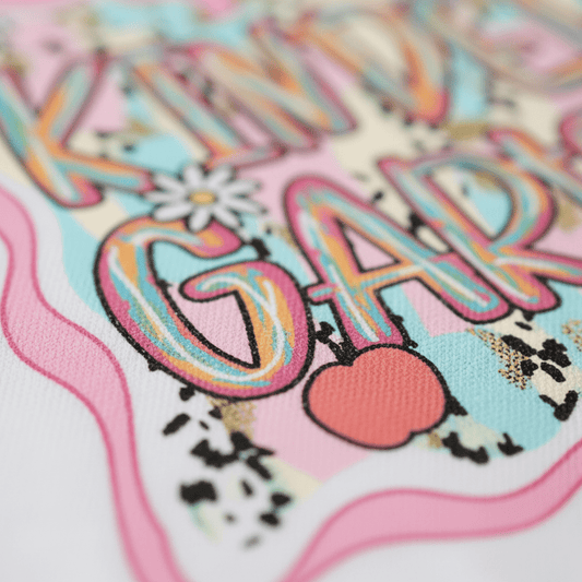Close-up of colorful, textured text reading KINDERGARTEN with playful patterns, pastel hues, floral accents, and a red apple illustration on a white background—perfect for the Toodleroos Kindergarten Youth Tee – Back to School Girls Shirt to celebrate the first day of school.