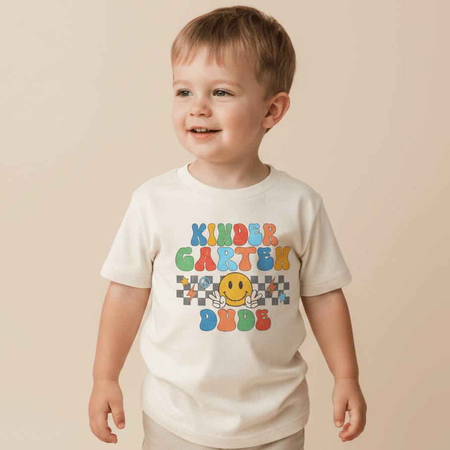 A young child smiles while wearing the Toodleroos Kindergarten Dude Youth Shirt – a retro back to school tee in white, featuring colorful text that reads “Kindergarten Dude” and a yellow smiley face graphic. The background is plain beige.