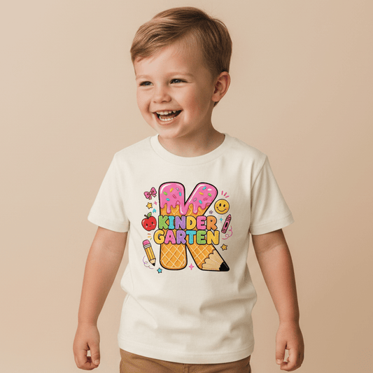 A cheerful young boy wears the Toodleroos Kindergarten Youth Girls Shirt – Ice Cream K Tee, featuring vibrant kindergarten-themed designs like pencils, ice cream, stars, and smiling faces—making it the ideal first day of school outfit as he stands against a beige background.