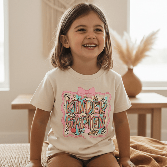A smiling young girl sits indoors wearing the Toodleroos Kindergarten Youth Tee – Back to School Girls Shirt, which features colorful “KINDERGARTEN” text adorned with bows and animal prints—ideal for first day of school or back to school outfits. Dried plants and a table appear softly blurred in the background.