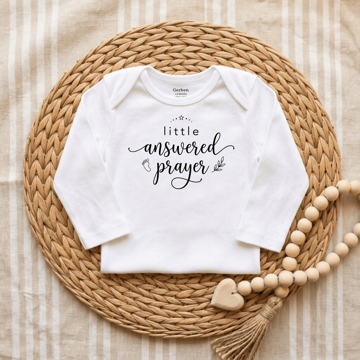A white Toodleroos Little Answered Prayer Onesie – Faith Baby Bodysuit with “little answered prayer” printed on the front is displayed on a round woven mat, beside a wooden bead garland featuring a heart and tassel—making it a perfect baptism gift or keepsake bodysuit for your precious little one.