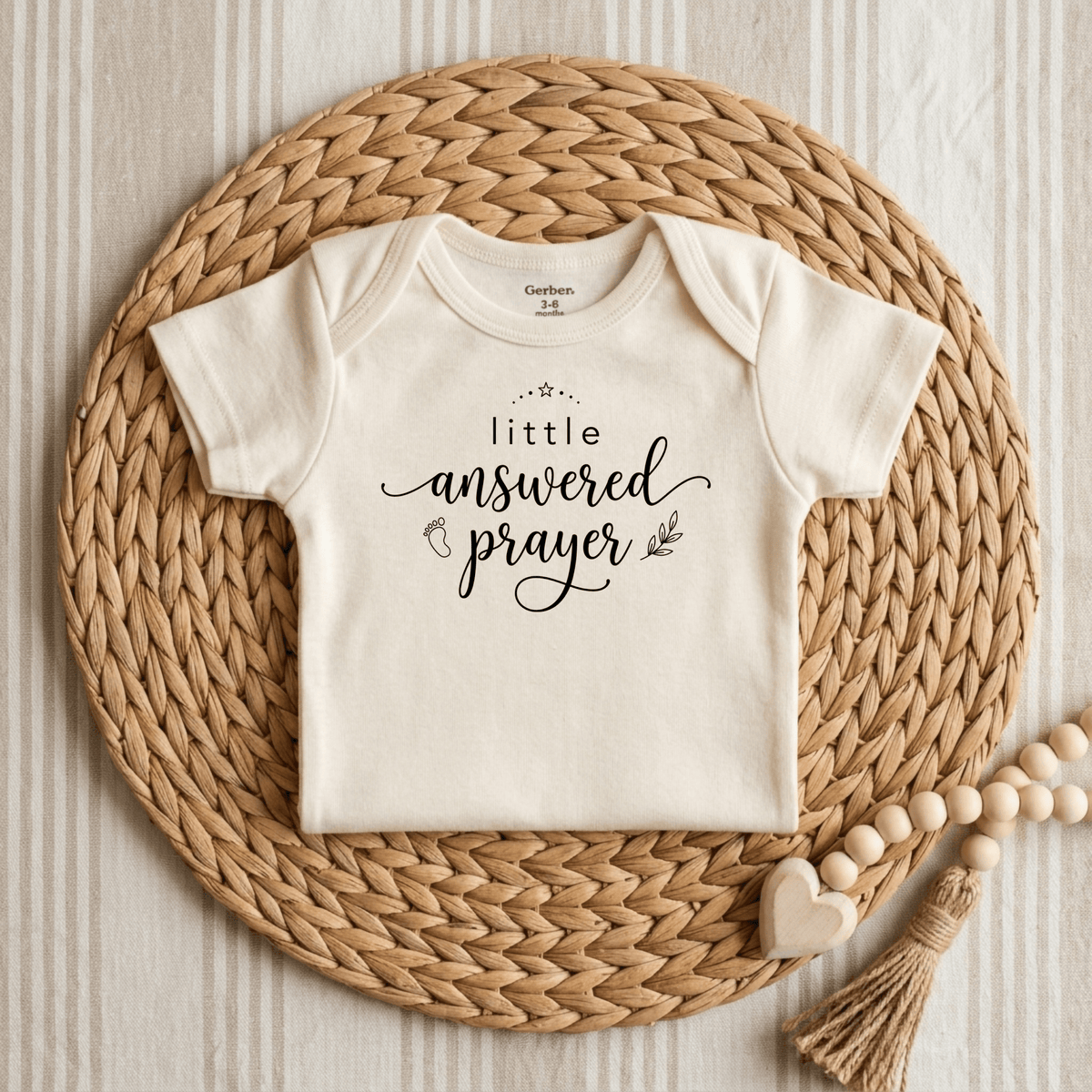 The Toodleroos “Little Answered Prayer Onesie – Faith Baby Bodysuit” features cream-colored Gerber short sleeves with elegant cursive lettering, shown atop a woven mat with a beaded tassel and heart accent on a striped backdrop—an ideal baptism gift for babies.