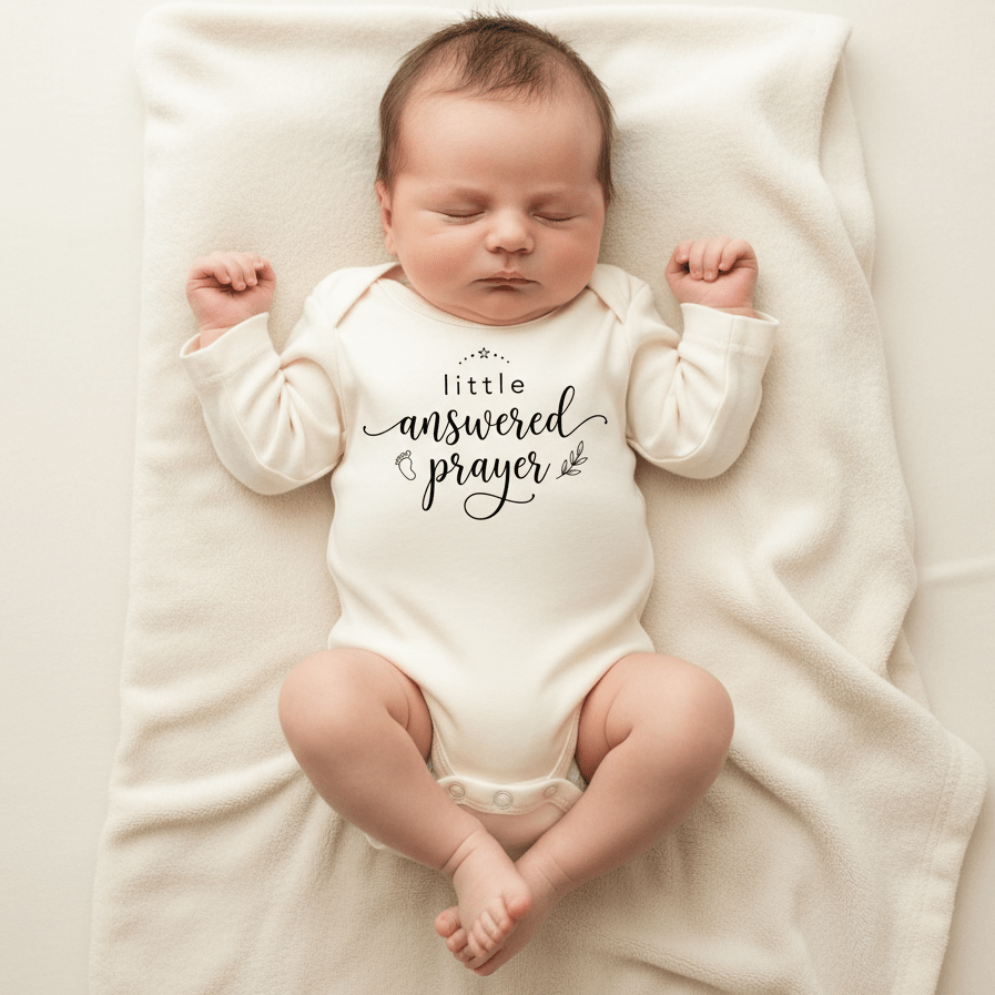 A sleeping baby lies on a white blanket, wearing a white Toodleroos Little Answered Prayer Onesie – Faith Baby Bodysuit, featuring the words “little answered prayer” in black script—a charming bodysuit that makes the perfect baptism gift for babies.