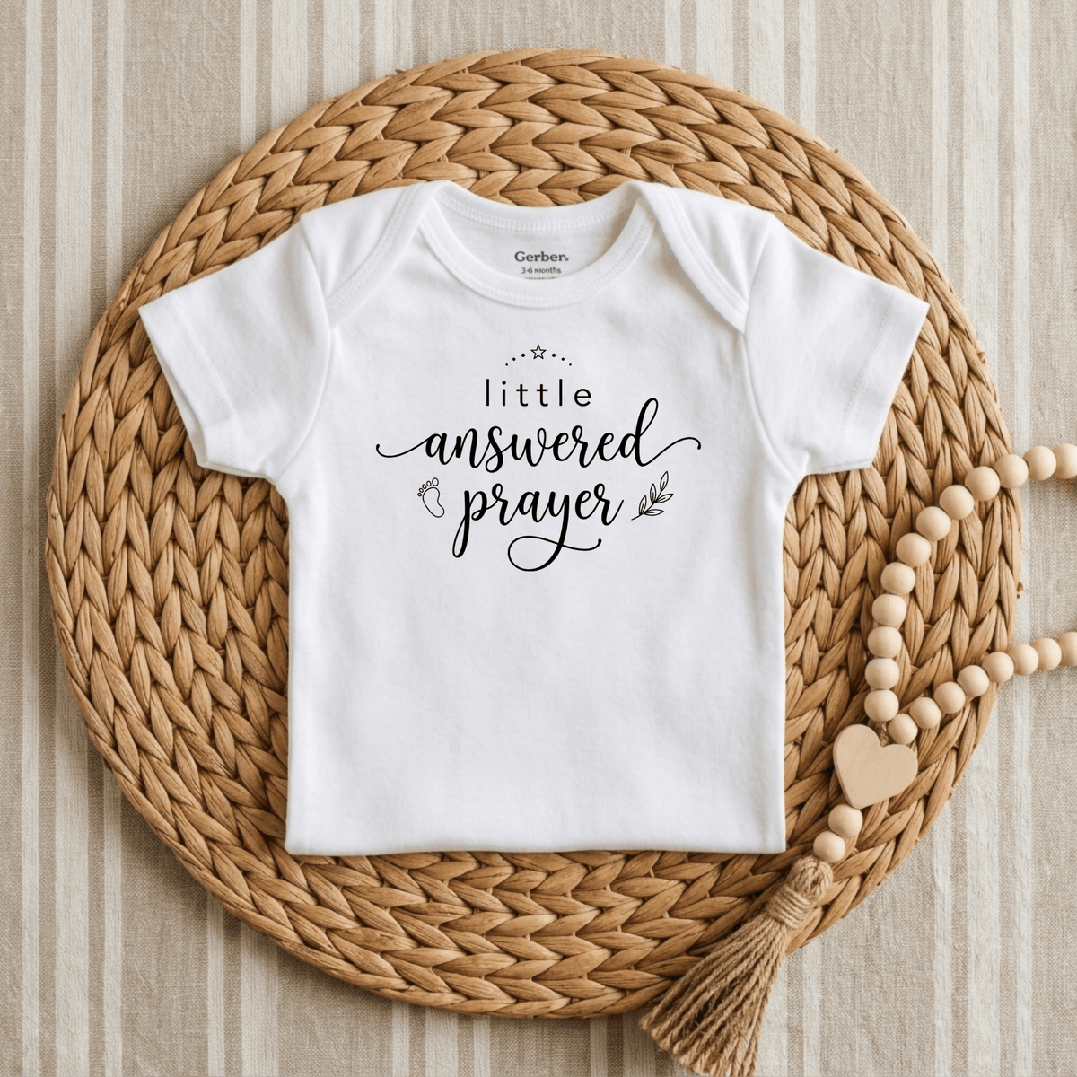 A white Toodleroos “Little Answered Prayer Onesie – Faith Baby Bodysuit,” featuring the phrase “little answered prayer” in black script, is displayed on a round woven placemat with a wooden bead garland beside it, all set against a striped fabric background. This Gerber short-sleeve bodysuit from Toodleroos makes a thoughtful baptism gift for babies.