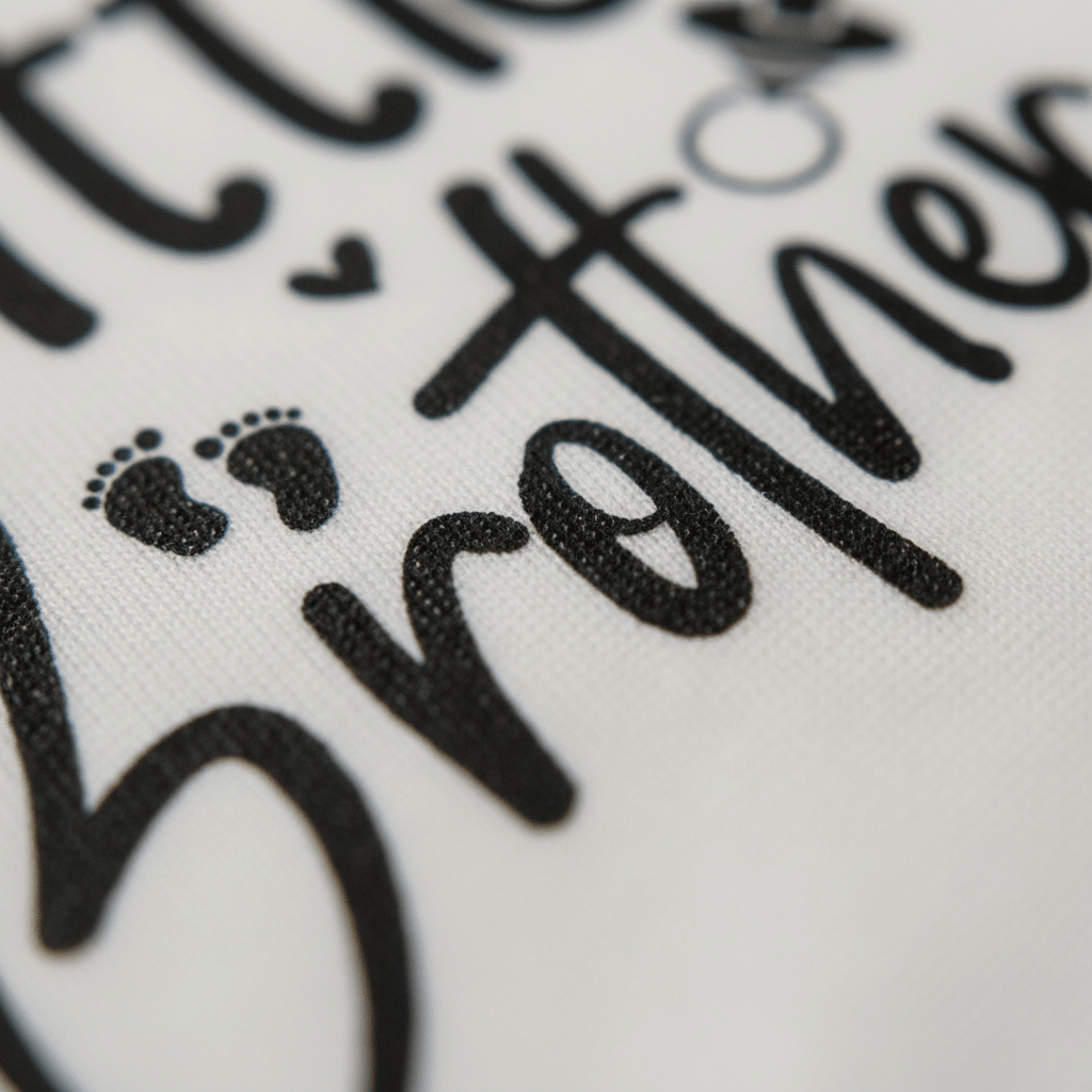 Close-up of fabric from the Toodleroos Little Brother Baby Onesie – Hand-Lettered Sibling Design, featuring “little brother” in black script with small hearts and a pair of baby footprints beside the text—a charming touch for a special coming home outfit or baby bodysuit.