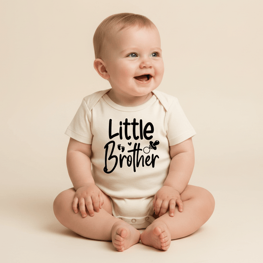 A smiling baby is seated on a plain light beige floor, wearing the Toodleroos Little Brother Onesie – Baby Boy Announcement Bodysuit. This white onesie, adorned with the words “Little Brother” and small heart graphics on the front, is perfect as a baby boy outfit or for a heartfelt pregnancy announcement.