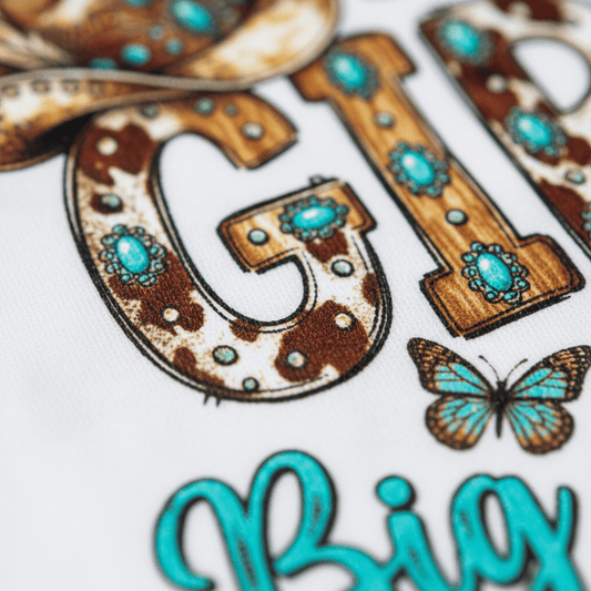 A close-up of the words “Little Girl Big Attitude” in cowboy-inspired lettering adorned with turquoise gemstones, brown spots, and a blue butterfly on white fabric—a playful detail from the Toodleroos Little Girl Big Attitude Kids Shirt – Western Cowgirl Tee. Perfect for any toddler cowgirl!.
