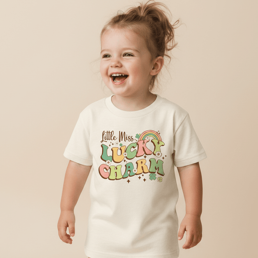 A smiling young child wears the Toodleroos Little Miss Lucky Charm Toddler Girls Shirt – St. Patrick’s Day Tee, featuring colorful text, pastel rainbow, clover, and star accents for a festive look. The soft beige background complements this adorable holiday outfit.