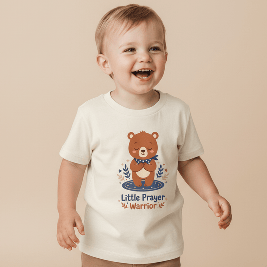 A cheerful toddler poses against a beige backdrop, wearing the Toodleroos Little Prayer Warrior Bear Toddler Shirt—a faith-filled cream t-shirt featuring an adorable bear illustration and “Little Prayer Warrior” text. This cute tee from Toodleroos is a perfect baptism gift for little ones learning about faith.