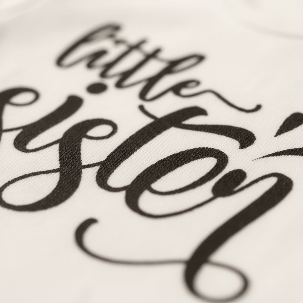 Close-up of hand-lettered black script reading “little sister” on white fabric, featured on the Toodleroos Little Sister Baby Onesie – Hand-Lettered Sibling Design, perfect for coordinating sibling outfits.