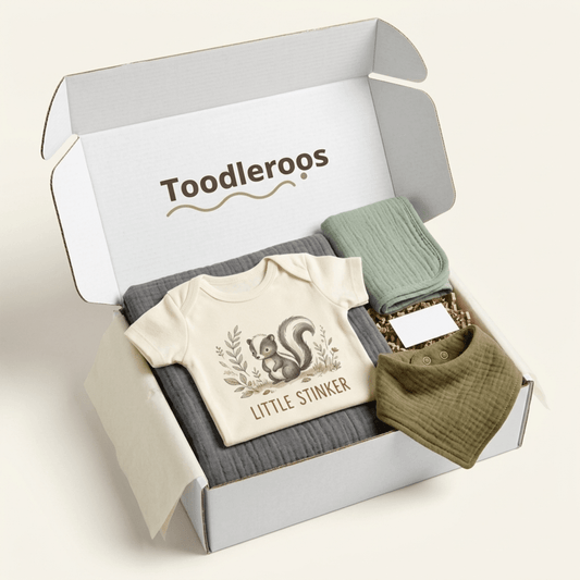 Open the Toodleroos Funny Baby Gift Box – Little Stinker Skunk Baby Shower Gift to reveal a neatly arranged cream baby shirt featuring a squirrel and “Little Stinker” text, a green muslin bib, a green swaddle, and a dark gray blanket—making it the perfect woodland-themed gift for any celebration.