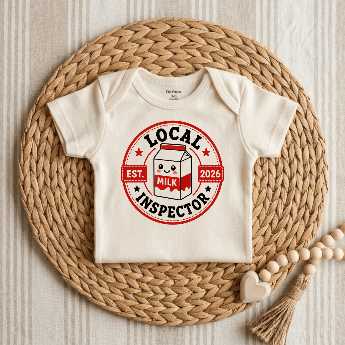 Dress your little one in the Toodleroos Local Milk Inspector Onesie – Funny Baby Bodysuit Est. 2026, a cream-colored unisex romper displayed on a woven placemat and adorned with a playful red and black “Local Milk Inspector” badge featuring an adorable milk carton and EST. 2026. Perfect for laughs, this cute baby onesie is complemented by wooden beads and a heart accent placed alongside for an extra charming touch!.