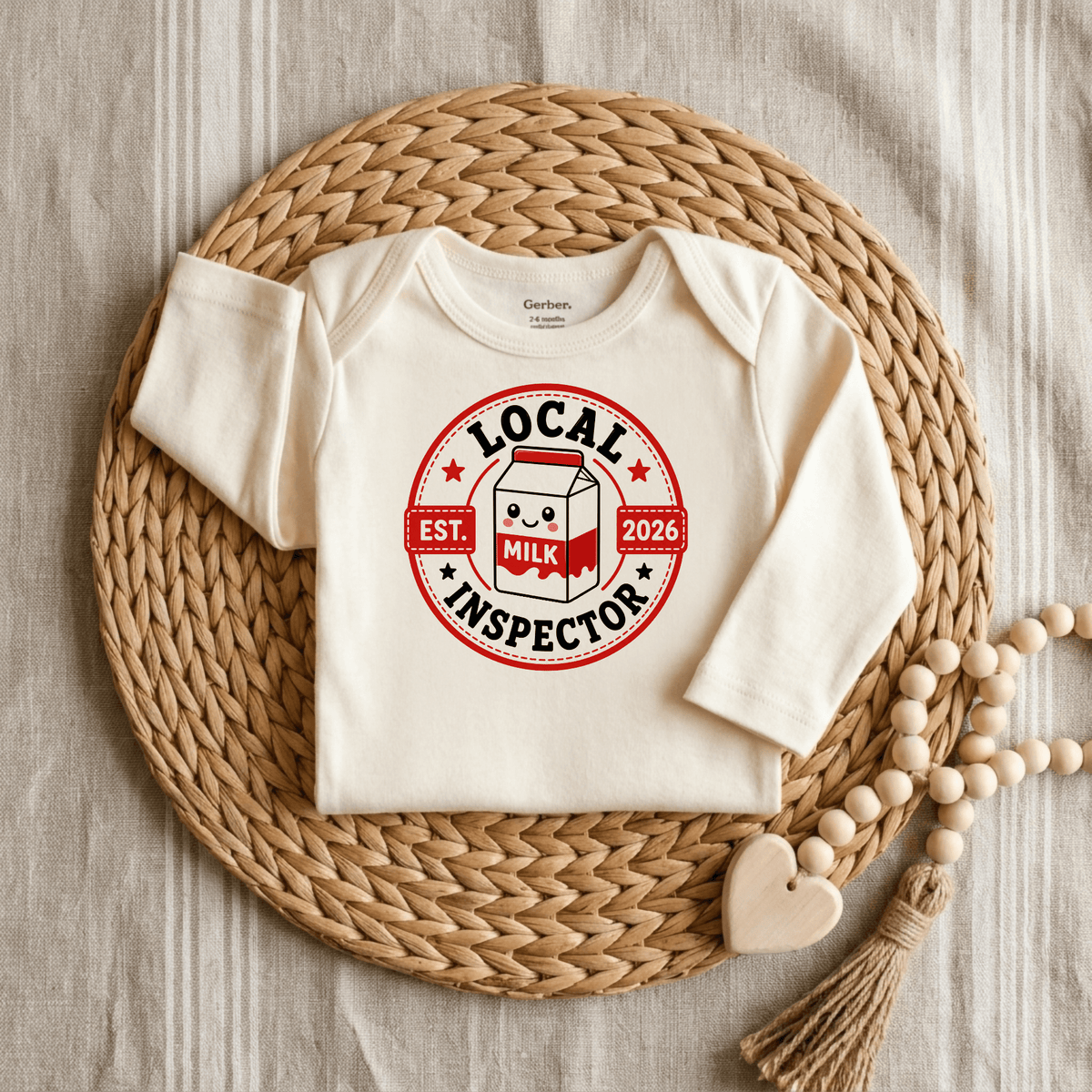 The Toodleroos Local Milk Inspector Onesie – Funny Baby Bodysuit Est. 2026 features a cream design with a red Local Milk Inspector badge and an adorable milk carton graphic. Displayed on a woven placemat alongside a wooden bead and heart teether, this stylish unisex baby onesie is perfect for anyone looking for funny baby bodysuits.