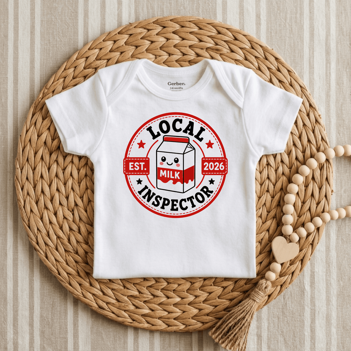 The Toodleroos Local Milk Inspector Onesie – Funny Baby Bodysuit Est. 2026 features a charming milk carton graphic and the playful text “Local Milk Inspector” alongside “Est. 2026.” Displayed on a woven mat with a beaded garland, this adorable unisex bodysuit is perfect for fans of funny baby onesies and makes an ideal baby gift.