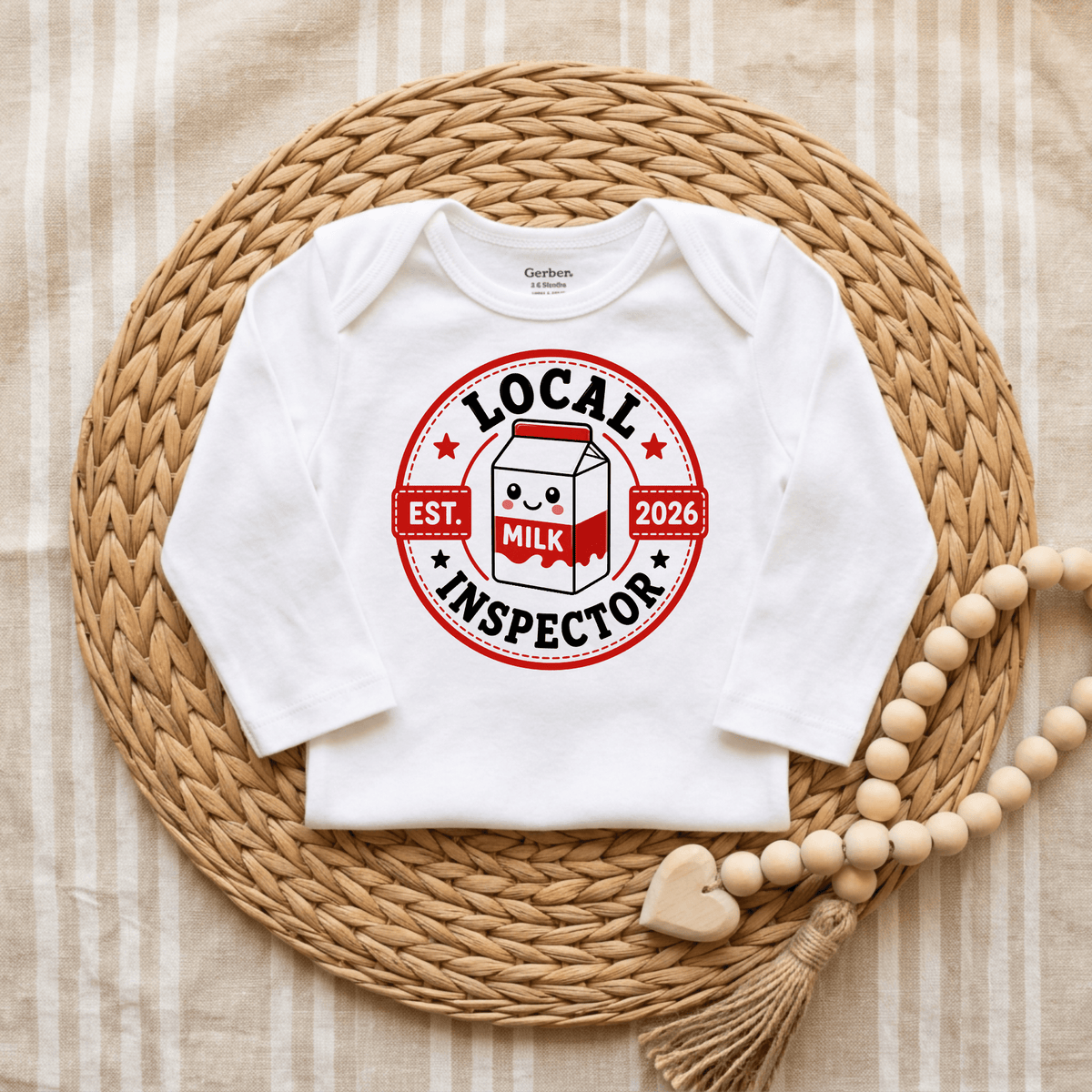 A long-sleeved white baby bodysuit from Toodleroos, the Local Milk Inspector Onesie – Funny Baby Bodysuit Est. 2026, is centered on a woven placemat. Its playful design showcases a red circle with “LOCAL INSPECTOR” and “EST. 2026” text, featuring a cheerful cartoon milk carton front and center—perfect for your little “milk inspector.” To the right sits a wooden bead garland, making this unisex onesie an irresistibly cute and funny choice for any baby. Add this whimsical Toodleroos piece to your collection