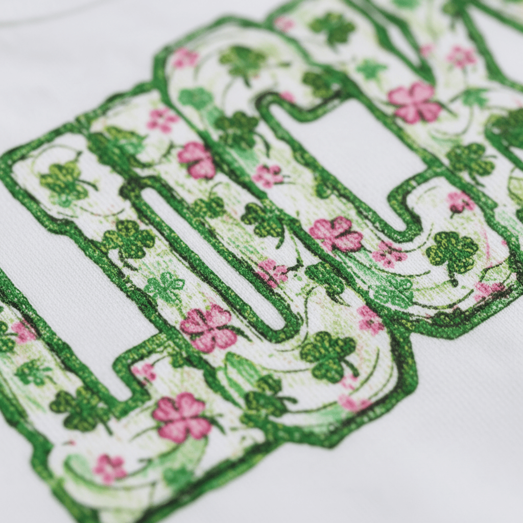 Close-up of the word LUCKY printed on soft fabric, with green clovers and pink flowers accenting the letters on a white background—an adorable choice for St. Patrick’s Day, featured on the Lucky Floral Clover Tee by Toodleroos.