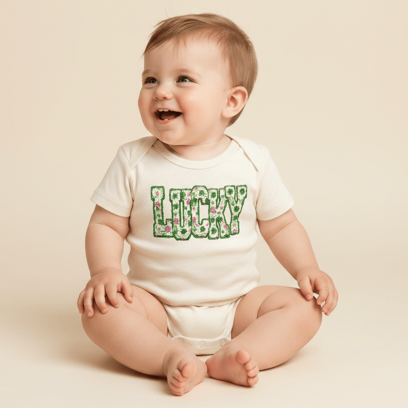A smiling baby sits on a light-colored background, wearing the Toodleroos “Lucky Floral Clover Tee”—a charming baby St. Patrick’s Day shirt featuring the word LUCKY in green floral letters, perfect for celebrating the festive occasion.