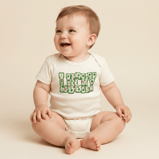 A smiling baby sits on a light-colored background, wearing the Toodleroos “Lucky Floral Clover Tee”—a charming baby St. Patrick’s Day shirt featuring the word LUCKY in green floral letters, perfect for celebrating the festive occasion.
