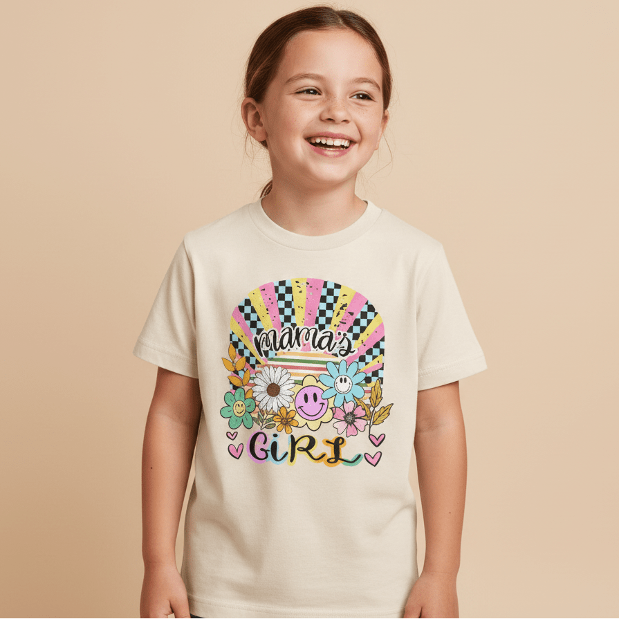 A smiling young girl wears the Toodleroos Mama’s Girl Retro Kids Shirt – a cream-colored boho floral tee featuring colorful “Mama’s Girl” text with flowers, hearts, and smiley faces on a pastel rainbow background.