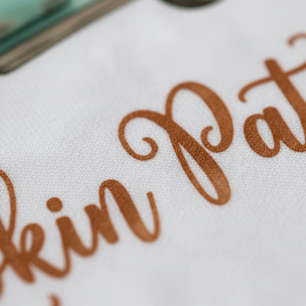 Close-up of brown cursive text reading “Meet Me at the Pumpkin Patch” printed on white fabric, with part of a green object visible in the corner—a cozy fall kids shirt from Toodleroos, perfect for autumn outings.