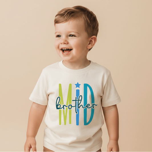 A smiling young boy stands against a beige background, wearing the Toodleroos Middle Brother Toddler Shirt – Colorful Sibling Design, featuring “MID brother” in bright blue and green letters—an ideal choice for a middle brother or sibling announcement.
